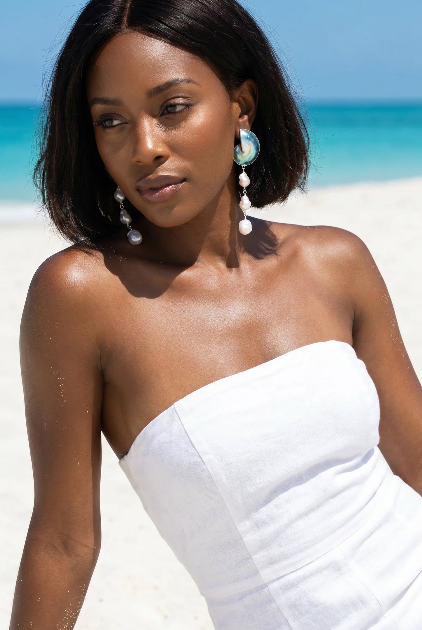 Woman in a white strapless dress wearing 925 silver encased nautilus shell cascade earrings on a beach with ocean backdrop | Lily Frank Jewelry
