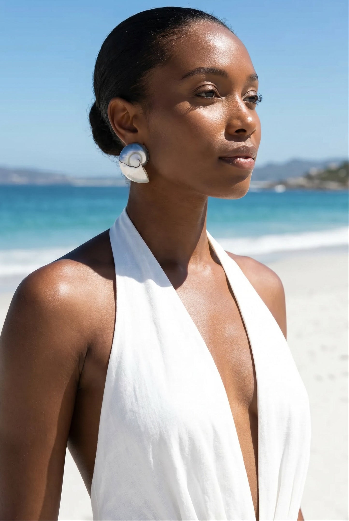 Woman in a white dress and a pair of  large nautilus shell drop earrings standing on a beach with clear blue sky and ocean | Lily Frank Jewelry