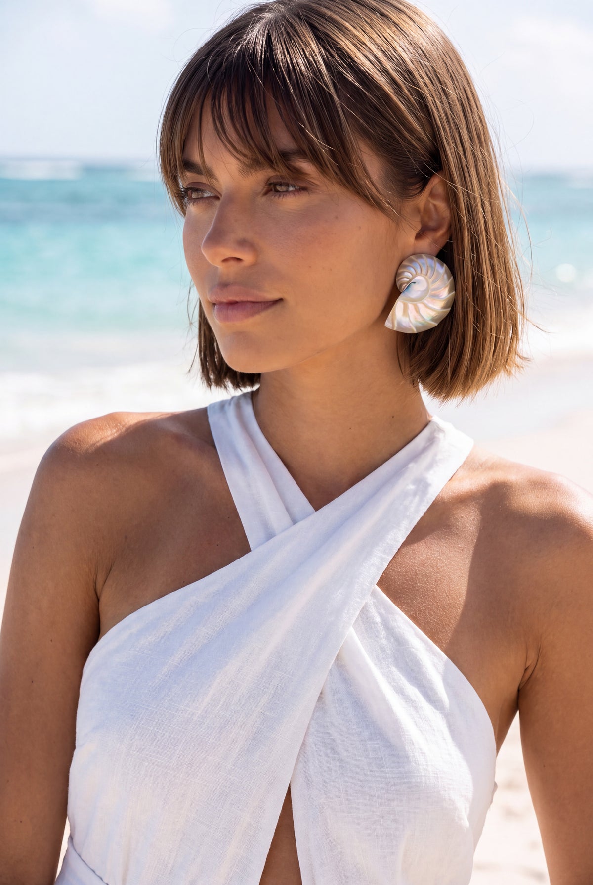 A woman with a short brown bob hairstyle stands on a beach wearing a white halter-style wrap dress highlgihted by a pair of large pearl nautilus shell eariings. The background shows a blurred turquoise ocean and white sand | Lily Frank Jewelry