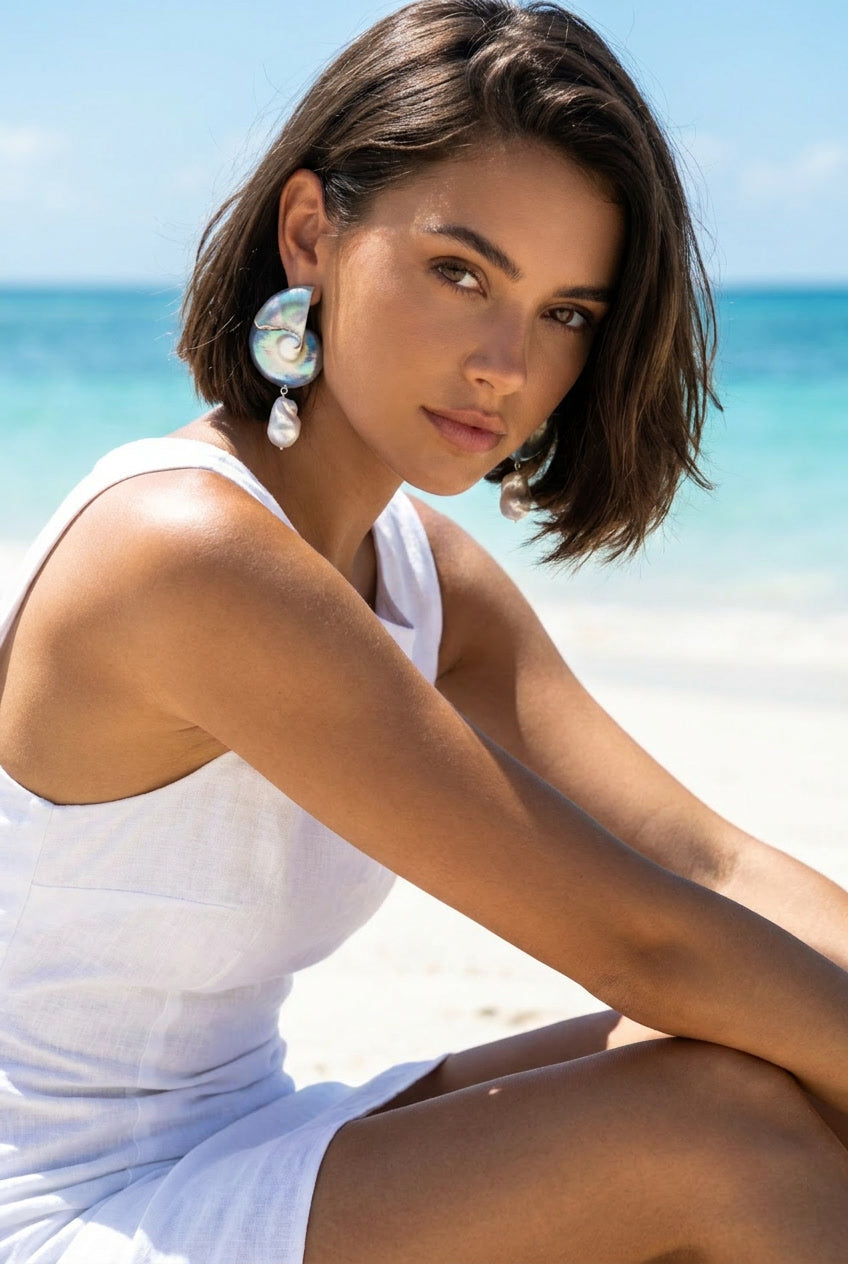 Woman sitting on beach wearing large two-toned nautilus shell drop earrings in white — Lily Frank Jewelry
