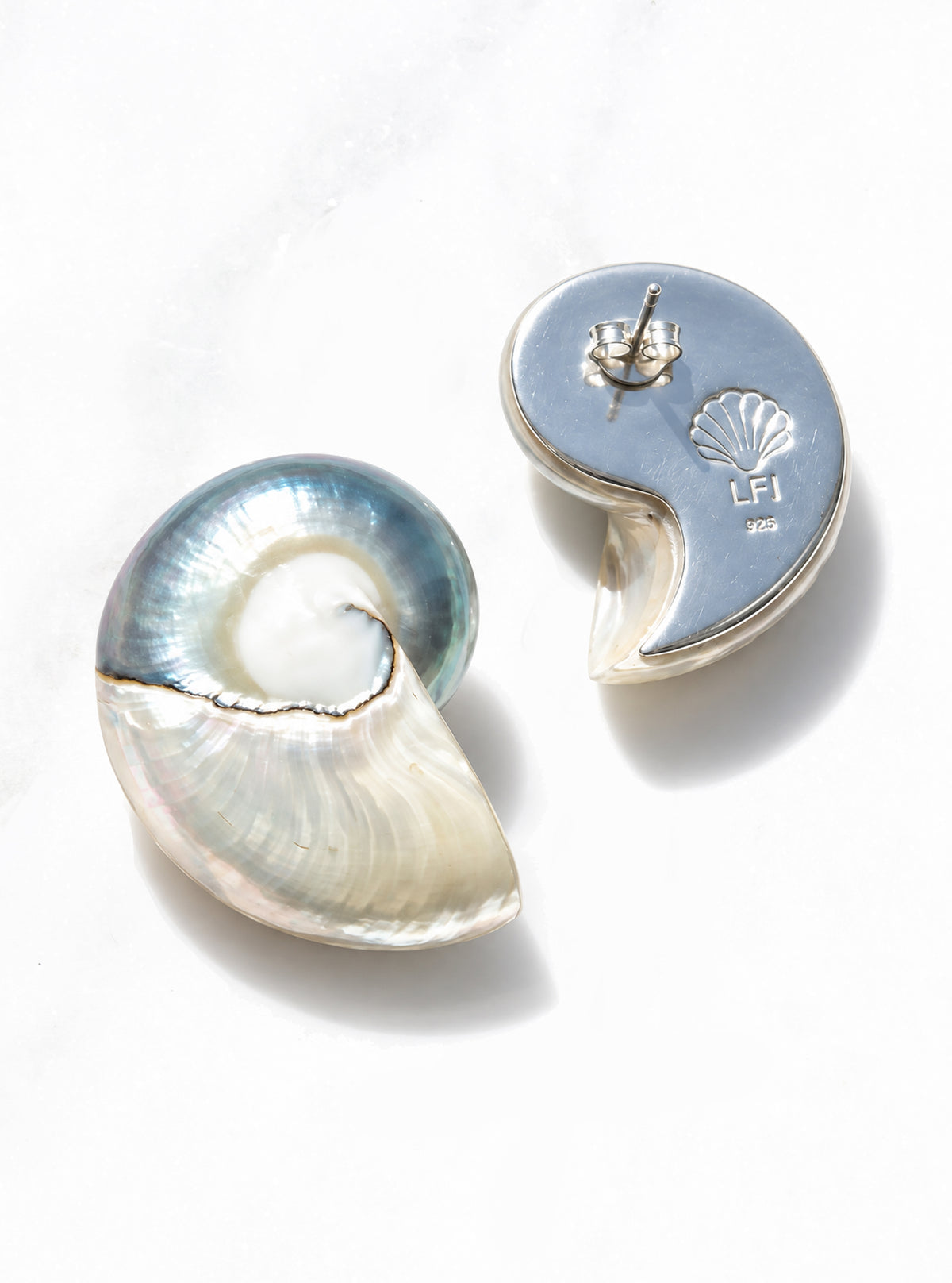 A pair of large two toned nautilus shell earrings displayed on a clean white surface. One earring is shown from the front, revealing a shimmering iridescent pearl interior, while the other is turned to show the polished 925 silver back with a shell engraving and post.