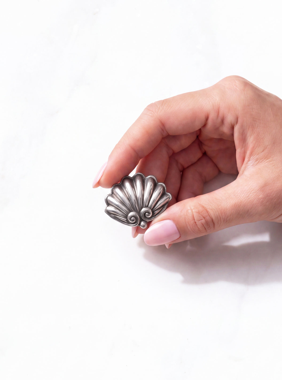 A hand with light pink manicured nails holds a vintage Margot de Taxco silver shell brooch against a clean white marble background | Lily Frank Jewelry