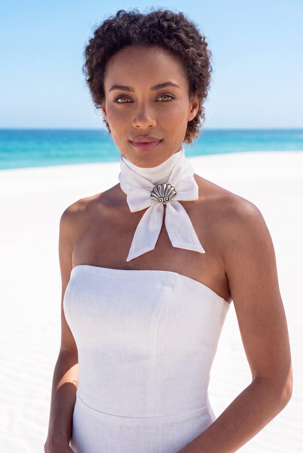 A woman with short curly dark hair stands on a white sand beach wearing a strapless white dress with a large white bow at the neck adorned with a vintage Margot de Taxco silver shell brooch | Lily Frank Jewelry