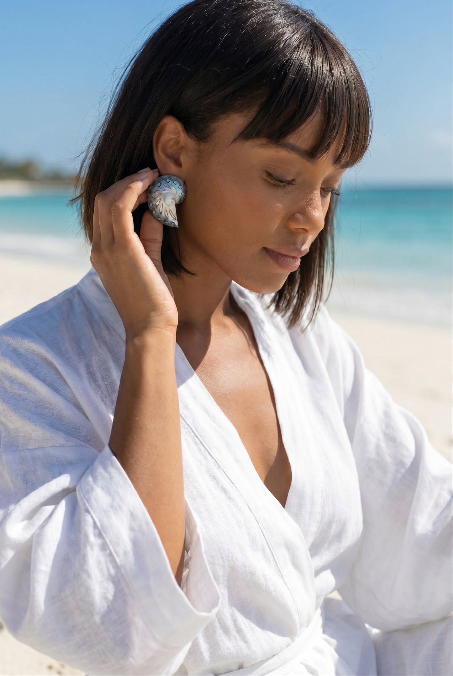 Woman on a beach wearing a white outfit and a pair of medium jaguar shell earrings adjusting the earrings | Lily Frank Jewelry