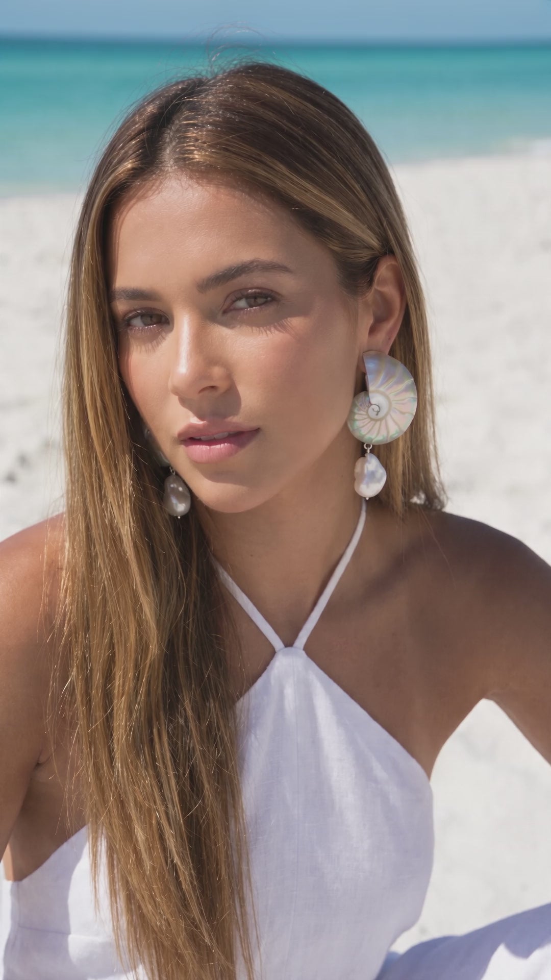 Video of a model wearing Large Pearl Nautilus Shell Drop Earrings with a white halter top on the beach | Lily Frank Jewelry