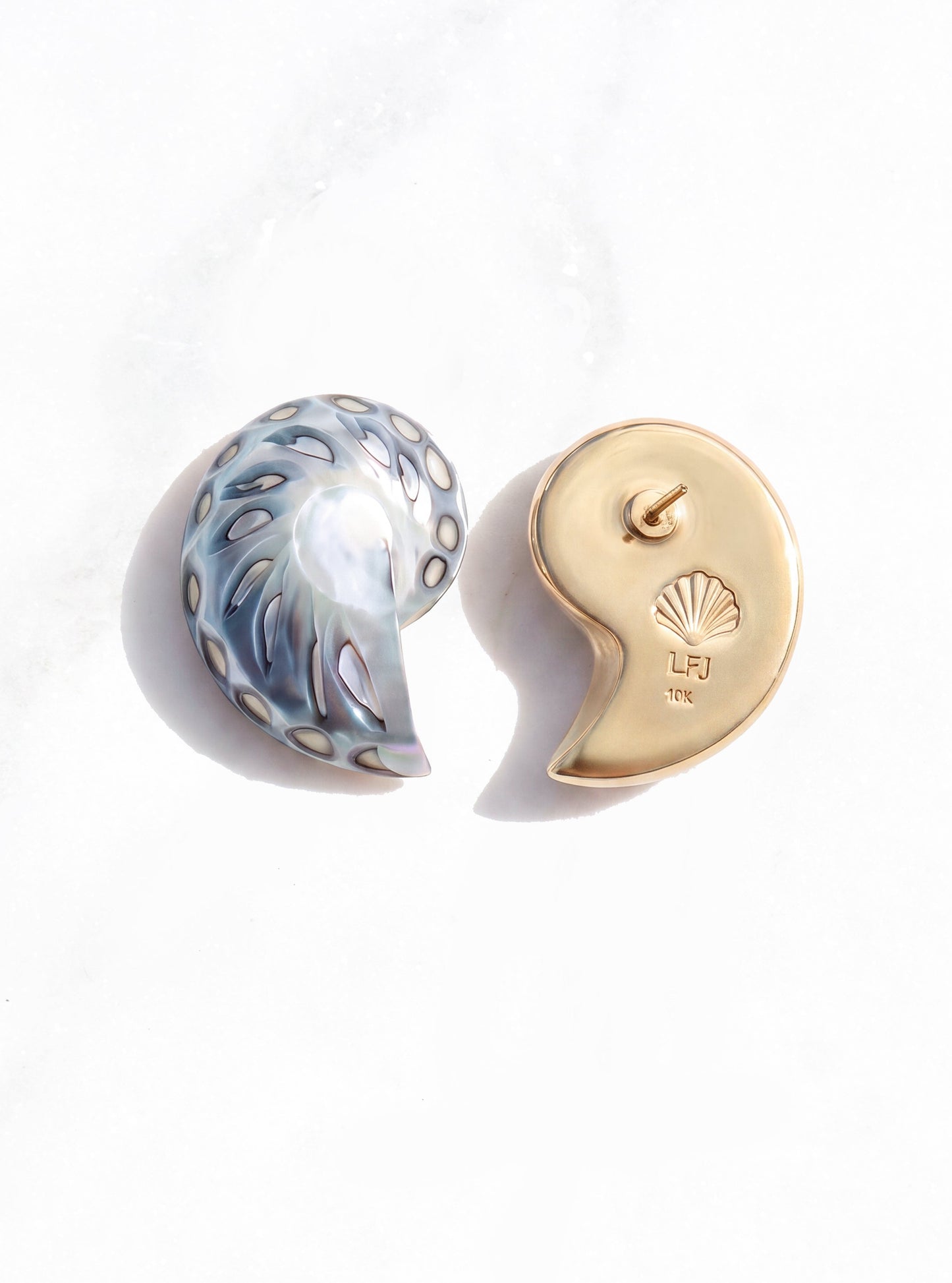 small jaguar shell earrings displayed on a white marble surface. One earring features a blue-gray and white marbled pattern, while the other is a polished 18k gold with the LFJ hallmark | Lily Frank Jewelry
