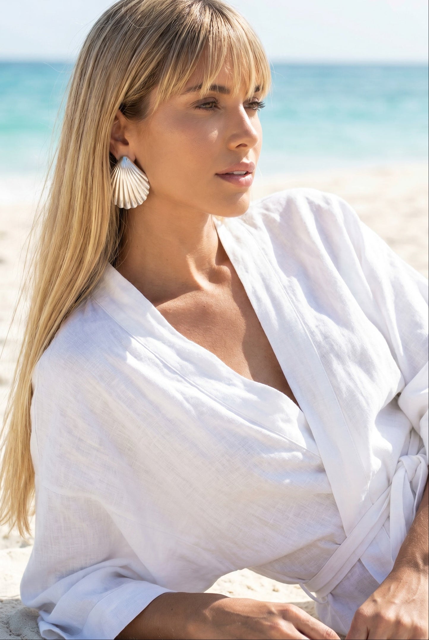 Woman in a white outfit wearing a pair vintage oversized 925 silver shell earrings sitting on a beach with ocean in the background | Lily Frank Jewelry 